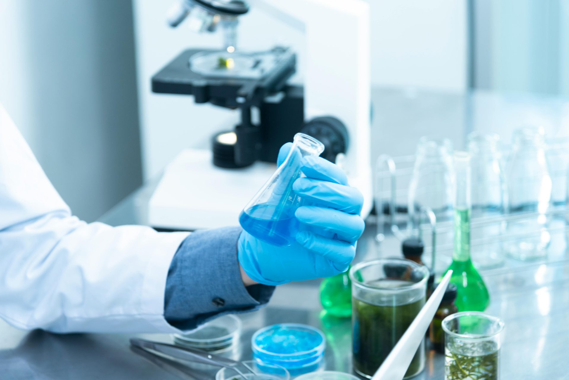 Wissenschaftler mit Handschuhen analysiert blaue Flüssigkeit in einer Laborumgebung mit Mikroskop und Glaswaren.